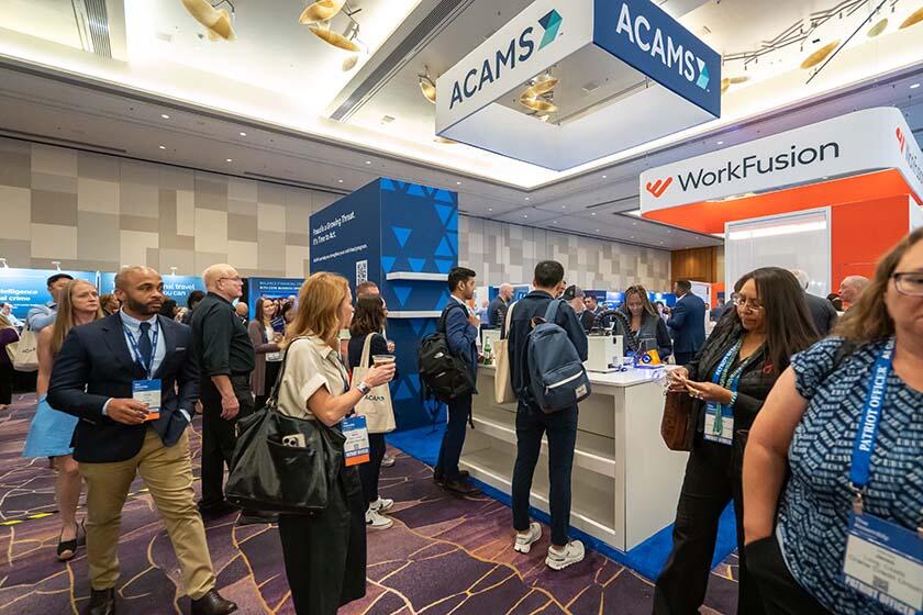 A diverse group of people stands in a convention hall surrounded by vibrant blue and orange booths.