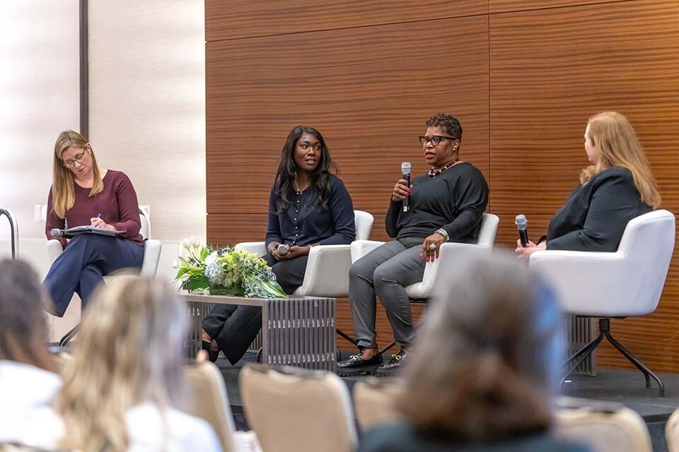 Four women seated in chairs, engaged in discussion at a professional conference setting.