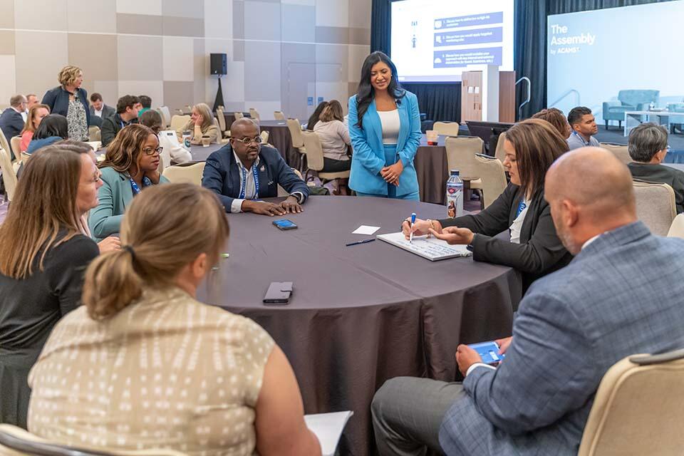 A diverse group of individuals engaged in discussion while seated at tables in a well-lit conference room.