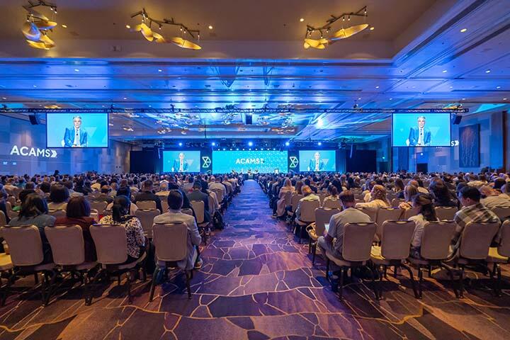 A spacious room crowded with attendees focused on a speaker delivering a presentation at the front.