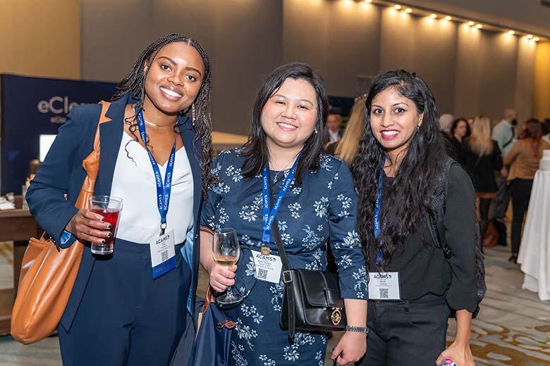 Three women at an event, smiling joyfully while holding drinks in their hands, creating a lively atmosphere.