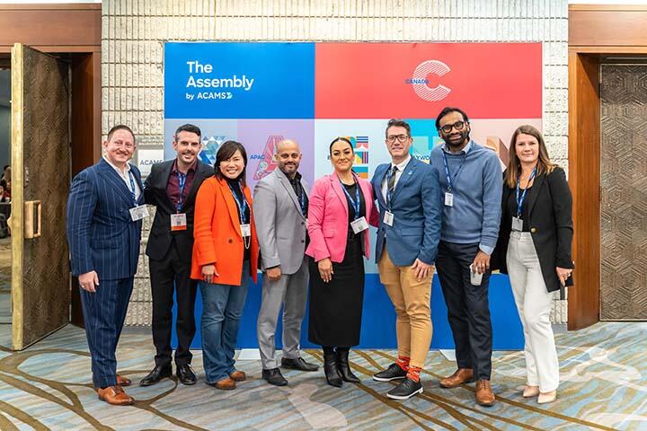 A diverse group of individuals stands together in front of a large banner, smiling and engaging with one another.